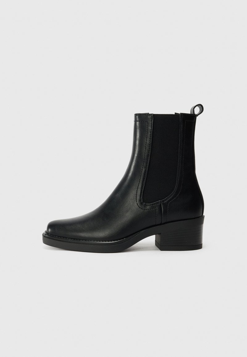 Bottine en cuir noir avec talon bloc, panneaux latéraux élastiques et languette à l’arrière sur fond gris clair.