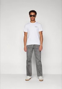 T-shirt branco de manga curta com um pequeno logótipo, combinado com calças cinzentas texturizadas e ténis brancos com solas castanho-claras. Óculos de sol pretos usados.