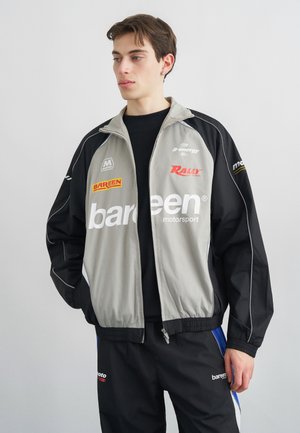 Jeune homme portant une veste de sport automobile noire et grise avec des logos et un pantalon noir assorti, debout devant un fond gris uni.
