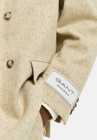 Manche de blazer en chevron beige avec des boutons marron et une étiquette de marque GANT, main dans la poche, poignet de chemise blanche visible.