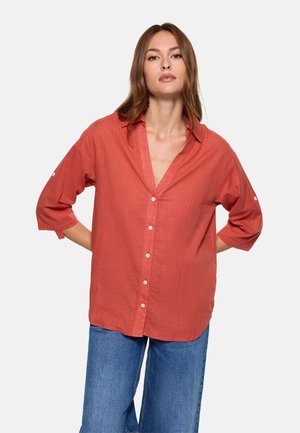 Mujer con camisa naranja de manga larga y botones, de corte holgado, y jeans azules, de pie con las manos detrás de la espalda frente a un fondo liso.