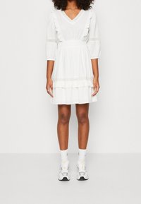 Mujer vestida con un vestido blanco de longitud a la rodilla, con detalles de volantes y adornos de encaje, combinado con calcetas blancas y zapatillas blancas, de pie frente a un fondo sencillo.