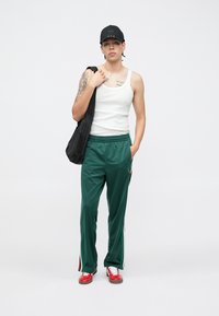 Πράσινο παντελόνι φόρμας από πολυεστέρα με ρίγες στο πλάι, συνδυασμένο με λευκό tank top. Μαύρο καπέλο και κόκκινα αθλητικά παπούτσια, κρατώντας μια μαύρη τσάντα ώμου.