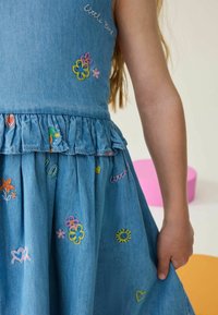 Džínová sukňa s rozšírenou siluetou. Má farebné vyšívané kvetinové a srdcové vzory. Volánový pás, svetlo modrá látka a bezrukávový dizajn.