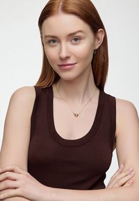 Złoty naszyjnik z wisiorkiem w kształcie serca na delikatnym łańcuszku, noszony na brązowej bluzce bez rękawów z dekoltem w serek. Gładkie, polerowane wykończenie metalu.
