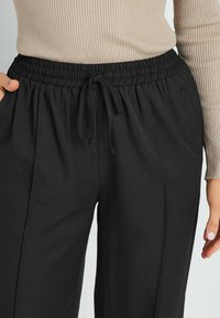 Zwarte elastische taillebroek met een trekkoord, geplooide voorkant en zijzakken. De stof heeft een zachte textuur.