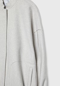 Giacca grigia chiara in misto lana con colletto rotondo, spalle scese, tasche laterali e chiusura frontale con zip pulita. Texture morbida e design minimale.