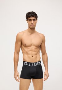 Crne bokserice s bijelim logom "CALVIN KLEIN" na pojasu. Glatka, rastezljiva tkanina. Model pozira bez majice.