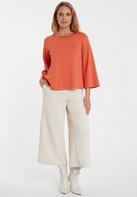 Oranges Langarmoberteil mit Rundhalsausschnitt, kombiniert mit cremefarbenen, weit geschnittenen, gesteppten Hosen und weißen, hohen Stiefeln, vor einem schlichten Hintergrund.