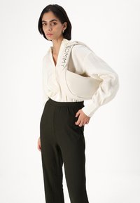 Cream-colored shoulder bag with a curved shape and embossed "TOMMY" logo on the strap. Model wears a white shirt and dark green pants.