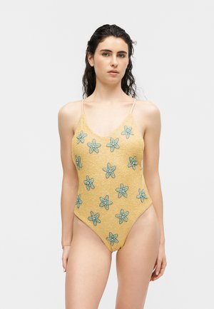 Tânără cu părul ud, purtând un costum de baie întreg galben decorat cu modele florale albastre pe un fundal gri deschis.