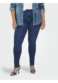 Vaqueros pitillo de tiro alto en azul oscuro hechos de mezclilla, combinados con una camisa clara de mezclilla. Los zapatos son tacones abiertos de color azul oscuro.