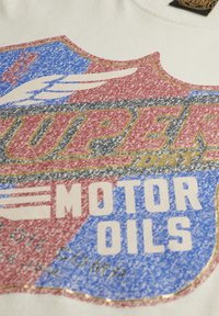 Camiseta de algodón de color claro con un diseño gráfico multicolor. Incluye texto en rojo, azul y blanco con una textura desgastada y acentos dorados.