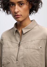 Béžová zipová košile s malým límečkem, dvěma kapsami na prsou a strukturovanou tkaninou. Obsahuje jemný šev přes ramena.