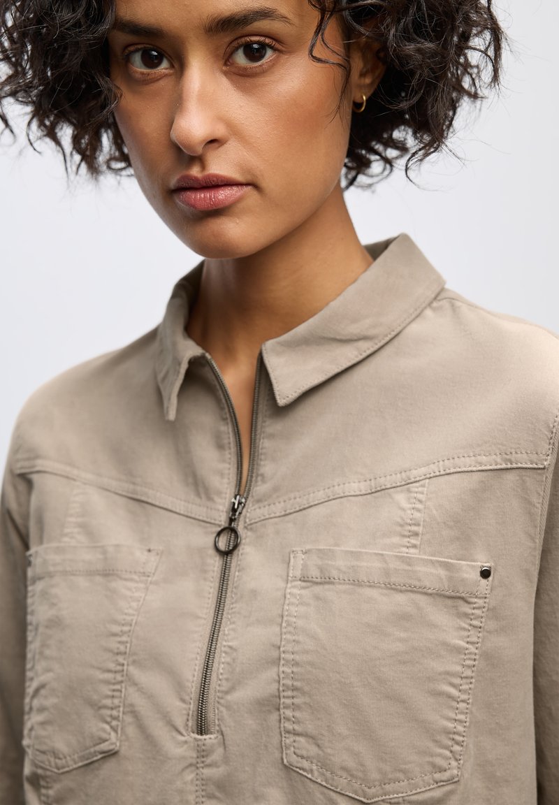 Vaahto beige zip-up-paita, jossa on pieni kaulus, kaksi rintataskua ja teksturoitu kangas. Ominaisuutena hienovarainen saumatieto hartioiden yli.