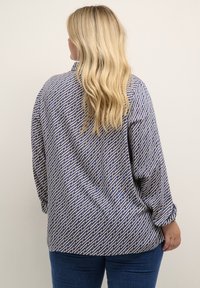 Femme blonde de dos, portant une chemise à manches longues bleu et beige à motifs et un jean bleu foncé, se tenant devant un arrière-plan uni.