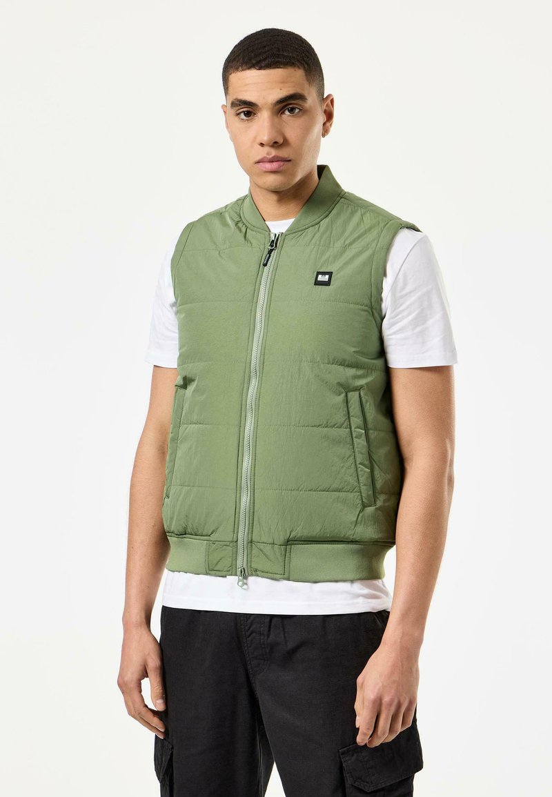 Grønn ermeløs vest med glidelås foran og sidelommer, med et lett, quiltet design og ribbestrikket kant. Brukt over en hvit skjorte.