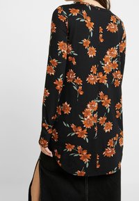 Mujer vestida con una blusa negra de manga larga con patrones florales en naranja óxido y una falda negra con una abertura lateral, vista desde atrás.