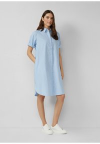 Donna in piedi che indossa un vestito camicia a righe verticali blu e bianche con maniche arrotolate e sneakers bianche su uno sfondo semplice.