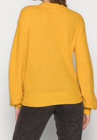 Femme portant un pull jaune ample et un pantalon sombre, vue de dos avec une main reposant sur le côté.