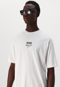 Valge puuvillane T-särk lühikestevarrukatega, millel on musta värvi trükitud logo tekst "DRYKORN FOR BEAUTIFUL PEOPLE" rinnal. Mustad ristkülikukujulised päikeseprillid.