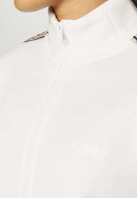 Hvit zip-up jakke med høy krage, glatt tekstur og subtil logo på forsiden. Inkluderer kontrasterende aksenter på skuldrene.