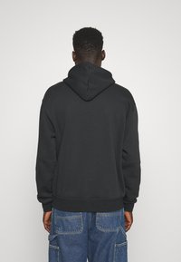 Uomo di spalle che indossa una felpa con cappuccio nera e pantaloni cargo blu in denim larghi su uno sfondo semplice.