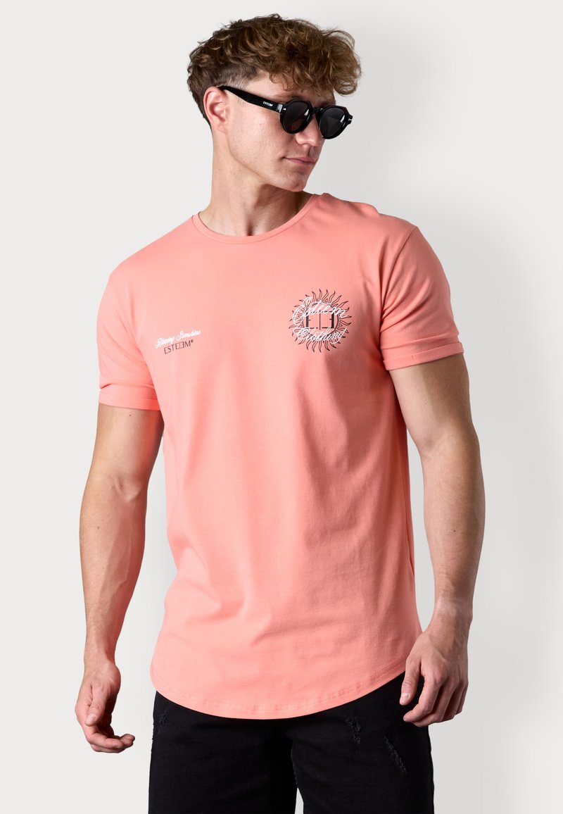 Coral cotton t-shirt with short sleeves, round neckline, and sun graphic detail on the chest, paired with black round sunglasses.
