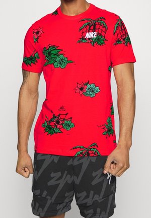 Mann trägt rotes Nike-T-Shirt mit grünen floralen und Netz-Designs, kombiniert mit schwarzen Shorts mit dunkelgrauen abstrakten Mustern.