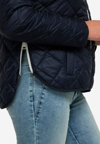 Marinblå quiltad jacka med en slät yta, som har vit dragkedja och sidofickor, tillsammans med ljusblå denimjeans.