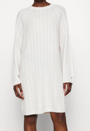 Personne portant une robe-pull blanche en maille torsadée à manches longues, col rond, poignets et ourlet côtelés.