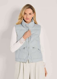 Helle blaue gesteppte Weste mit hohem Kragen, Druckknöpfen und zwei Fronttaschen, kombiniert mit einem weißen Langarmoberteil mit Spitzenakzenten.