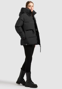 Crna puffer jakna s visokim ovratnikom i podesivom kapuljačom. Ima elastične manžete, prednji zip i dug, kutijasti oblik. Uparena s crnim tajicama i čizmama.