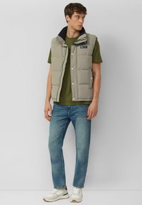 Gewatteerd mouwloos vest in lichtgrijs met zwarte kraag, gecombineerd met een olijfgroen T-shirt en lichtblauwe jeans, met casual sneakers.