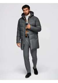 Cappotto imbottito grigio con cappuccio, dotato di zip frontale, indossato sopra una camicia a scacchi, abbinato a pantaloni grigi e stivali neri.