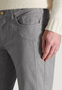 Pantalones de algodón grises con corte recto, costuras visibles, un bolsillo delantero con logo y cierre de botón. Detalle de tela texturizada.