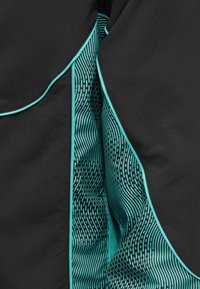 Tissu noir avec passepoil turquoise et bord incurvé, dévoilant partiellement un textile turquoise avec des motifs de lignes ondulées noires et de points.