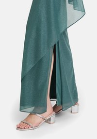 Turkusowa, błyszcząca sukienka o warstwowej konstrukcji, z wysokim rozcięciem na udzie. Srebrne sandały na słupku z cienkimi ramiączkami wysadzanymi kryształkami.