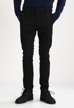 Mężczyzna ubrany w czarne spodnie o dopasowanym kroju, czarne sznurowane buty oraz ciemnoniebieską koszulę z długim rękawem, stojący z rękami w kieszeniach.