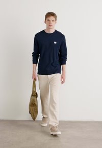 Donkerblauwe langetermijnsleeve shirt met een klein logo, beige broek en lichtbruine schoenen. Houdt een kaki tas vast, terwijl hij loopt op een grijze vloer tegen een witte muur.