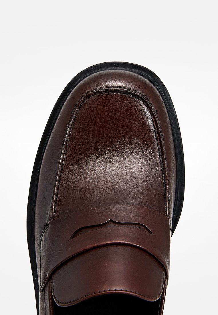 Mocassin en cuir marron avec un bout arrondi, des coutures prononcées, un détail découpé décoratif sur le coup de pied et une semelle en caoutchouc noire.