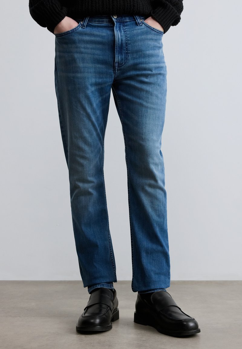 Ανοιχτό μπλε τζιν με ευθεία γραμμή, με μπροστινές τσέπες, απαλή ξεβάμματα και συνδυασμένα με μαύρα παπούτσια τύπου slip-on.