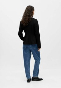 Melnas uzvalku žaketes ar strukturētām šuvēm, kas valkāta virs brīviem zilu džinsu biksēm. Melnas kurpes papildina tērpu. Aizmugures skats, vienkāršs fons.