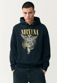 Melnā kapuce ar centrālu grafisko attēlu, kurā attēlots eņģelis ar spārniem, dzelteniem traipiem un virsū ar tekstu "NIRVANA".