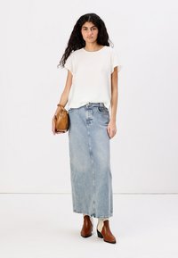 Top bianco a maniche corte, gonna maxi in denim azzurro chiaro, stivaletti marroni alla caviglia, che tiene una piccola borsa beige, esposto su uno sfondo semplice.