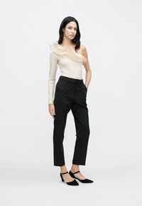 Smėlio spalvos vieno peties palaidinė su raukiniais, derinama su juodomis klasikinėmis kelnėmis ir juodais aukštakulniais su kulkšnies dirželiais, ant balto fono.