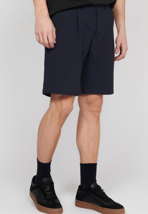Pantaloni scurți texturati de culoare albastru marin, cu un brâu elastic, prezentând pliuri frontale. Asortați cu adidași negri și șosete închise la culoare.