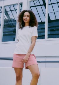 T-shirt bianco a maniche corte abbinato a pantaloni corti rosa con texture, caratterizzato da un collo quadrato e una piccola etichetta sulla tasca dei pantaloni.