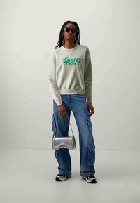 Szürke pulóver zöld "Sports Club" grafikával, kék cargo farmernadrág, ezüst válltáska és fekete napszemüveg. Texturált anyag és laza fazon.