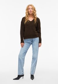 Maglione marrone a maglia strutturata con scollo a V, abbinato a jeans azzurri a gamba dritta e tacchi neri con punta affilata.